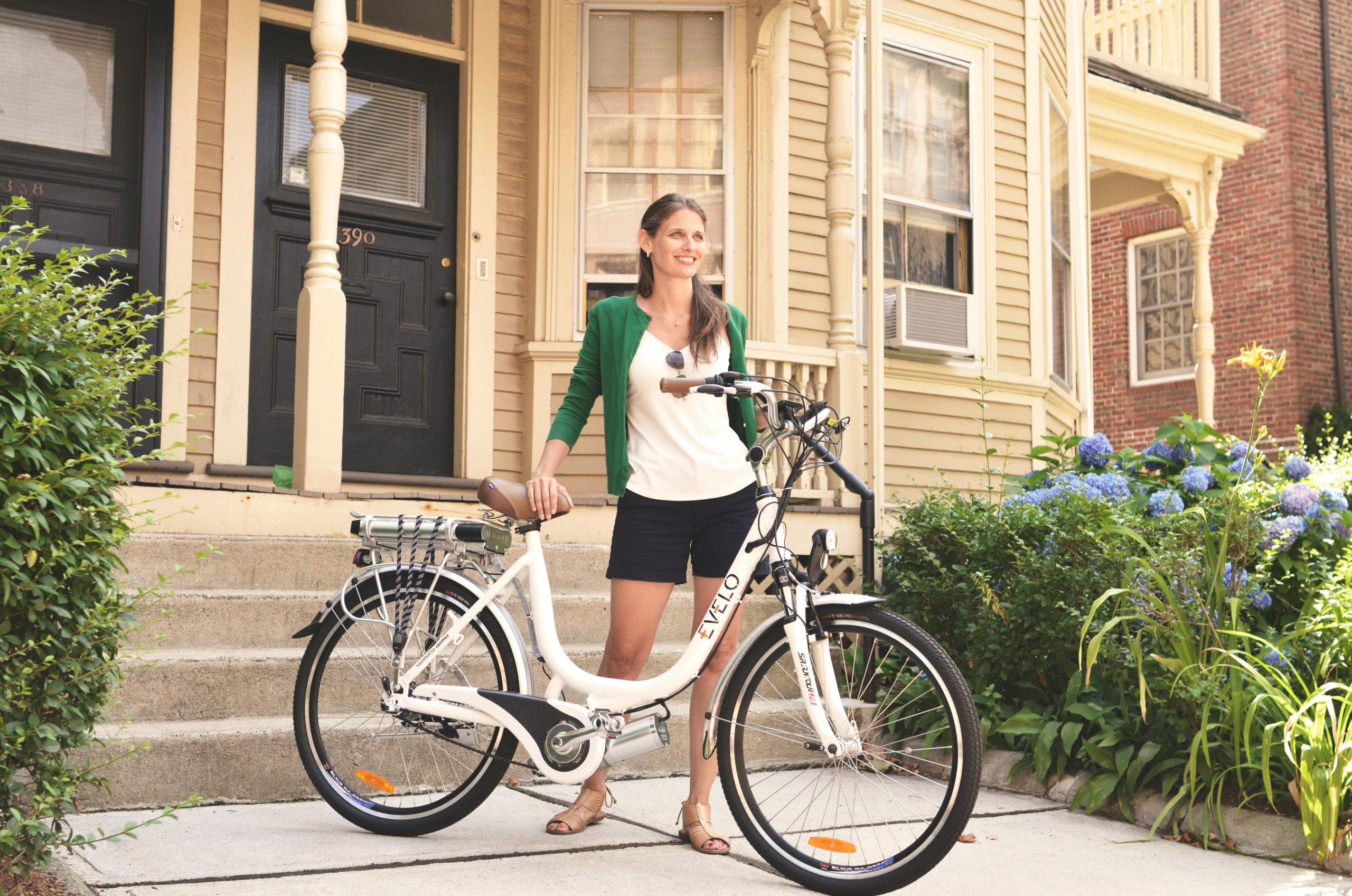 Woman with an electric bicycle in front of a suburban home, suggesting eco-friendly lifestyle and urban commuting.